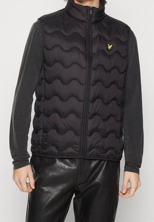 Mann trägt eine schwarze, wellenförmige, gesteppte Weste mit Reißverschluss über einem dunklen Langarmshirt und schwarzen Lederhosen, steht mit entspannten Armen.