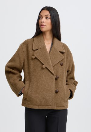 Femme aux longs cheveux raides portant un manteau en laine marron à double boutonnage, les mains dans les poches, regardant sur le côté sur fond blanc.