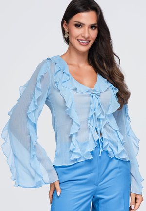 Femme souriante, portant un chemisier bleu clair transparent avec des volants et un nœud à l'avant, assorti à un pantalon bleu assorti, sur un fond clair uni.