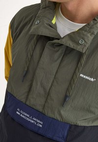 Chaqueta con capucha verde y amarilla que presenta un cordón de ajuste, paneles en contraste en azul marino y negro, y detalle del logo en el bolsillo delantero.