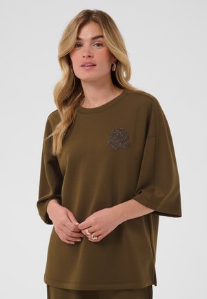 Mujer con el pelo largo y rubio, vistiendo una camiseta oversize verde oliva con un parche decorativo en el pecho, de pie frente a un fondo liso.
