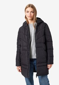 Doudoune noire avec capuche, présentant un design matelassé, une fermeture éclair et des poignets élastiques, portée sur un sweat-shirt gris et un jean bleu.