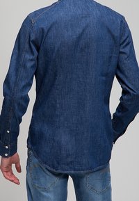 Homme portant une chemise en denim bleu foncé à manches longues et un jean bleu, de dos, les bras détendus sur les côtés.
