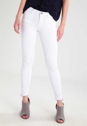 Femme portant un pantalon blanc slim avec des fermetures éclair aux chevilles, un haut bleu marine et des bottines ouvertes grises à talons, debout devant un fond uni.