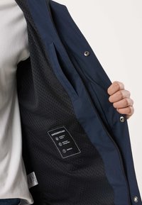 Ręka trzymająca otwartą granatową kurtkę, ukazującą czarną przewiewną siateczkową podszewkę z metką Swedemount, na której zaznaczone są właściwości wodoodporne, oddychające i wiatroodporne.