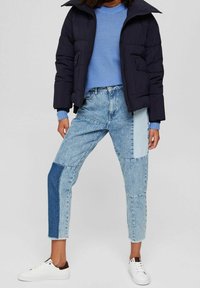 Kvinde iført en sort boblejakke, blå sweater, patchwork lys- og mørkeblå jeans samt hvide sneakers, stående med benene fra hinanden.