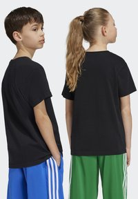 T-shirt en coton noir à manches courtes, avec un col rond. Deux enfants le portent, associé à des shorts de sport de couleurs contrastées.