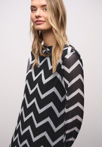 Černobílá halenka s cikcak vzorem, vysokým límcem a dlouhými průsvitnými rukávy, s texturovaným povrchem tkaniny.