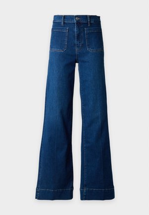 Femme aux longs cheveux noirs portant une chemise noire à fleurs boutonnée, un jean bleu à jambes larges et des chaussures noires à talons hauts, debout devant un fond uni.