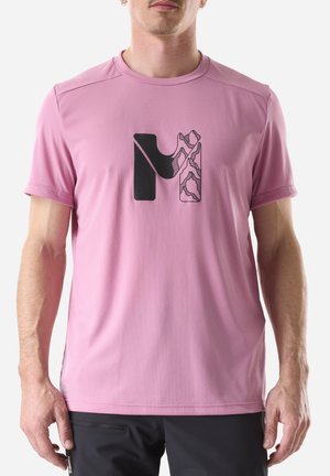 Homme portant une chemise à manches courtes rose avec un logo "M" stylisé en noir et à motifs, associée à un pantalon noir.