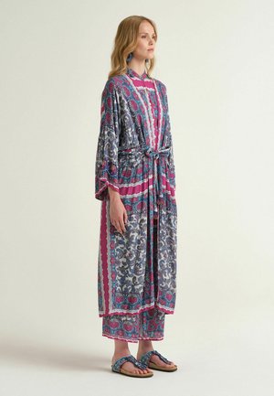 Donna in piedi con una lunga vestaglia a motivi con cintura annodata, pantaloni larghi abbinati e sandali slip-on decorati, su sfondo neutro.