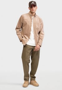 Man standing wearing brown baseball cap, beige button-up shirt over white tee, olive pants, and tan sneakers on plain background.