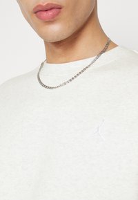 Närbild av en person som bär ett silverfärgat kedjehalsband och en ljuströja med en diskret Jumpman-logotyp på bröstet.