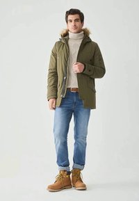 Giacca parka verde oliva con cappuccio rifinito in pelliccia, abbinata a un maglione di cashmere crema, jeans blu e stivaletti da lavoro in cuoio beige.