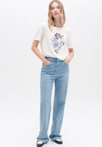 Weißes T-Shirt mit floralen Stickereien in Blau, Lila und Orange. Hellblaue, hochtaillierte Jeans mit entspannter Passform und geradem Bein.