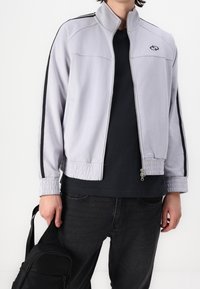 Lichtgrijze ritsjack met zwarte accenten, elastische manchetten en zoom. Draagt over een donker shirt, in combinatie met zwarte jeans en een zwarte tas.