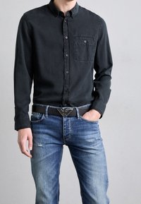 Man som bär en svart knappad skjorta, blå urblekta jeans med lätt nötning och ett svart bälte med en silverfärgad örnformad spänne.