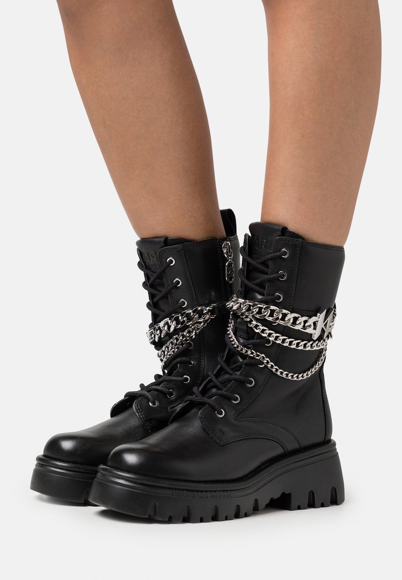 Schwarze Lederkampfstiefel mit dicken Sohlen, Schnürsenkeln, Reißverschluss an den Seiten und mehreren silbernen Kettenverzierungen um die Knöchel.