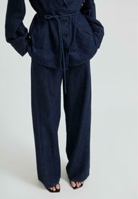 Temno modra denim obleka, ki vključuje suknjič s pasom in žepi ter široke hlače. Tkanina ima subtilno teksturo in globoko modro barvo.