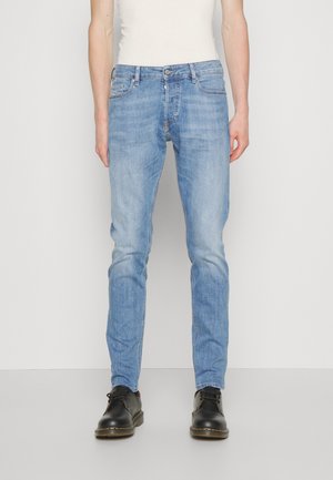 Mand iført lyseblå slim-fit jeans, sorte snøresko og en hvid ærmeløs skjorte stående mod en plain hvid baggrund.