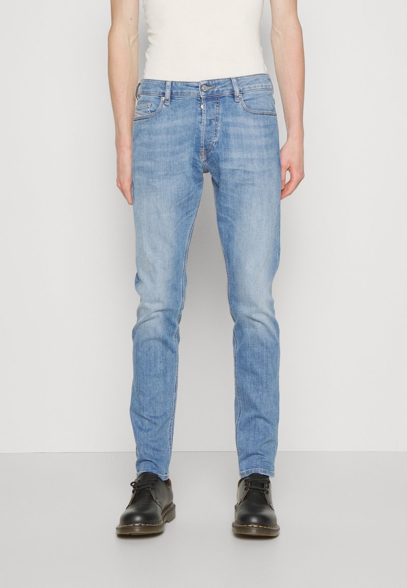 Homme portant un jean slim bleu clair, des chaussures noires à lacets et un débardeur blanc, debout devant un fond blanc uni.