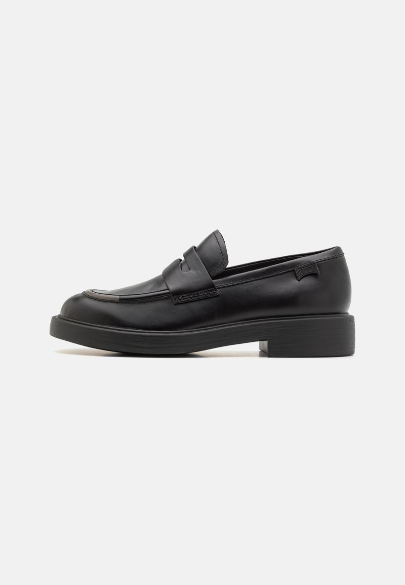 Mocassins en cuir noir avec un bout arrondi, des détails cousus et un petit talon carré. Ils possèdent une sangle à l'avant et un logo embossé sur le talon.