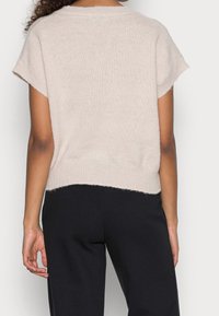 Lys beige sweater med korte ærmer, lavet af blødt, struktureret materiale. Afsluttet med en ribkant, båret over sorte bukser.