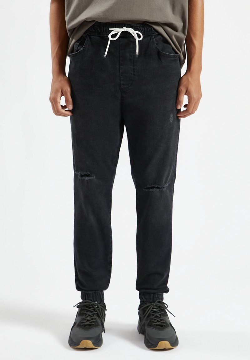 Czarne spodnie dresowe z elastycznym pasem i białym sznurkiem. Posiadają wyblakłe miejsca i przetarcia na kolanach. Gładka faktura denimu.