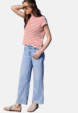 Frau mit Sonnenbrille, rot-weiß gestreiftem kurzärmligem Shirt, hellblauen weiten Jeans und beigen Sneakern, steht mit einer Hand am Kopf.