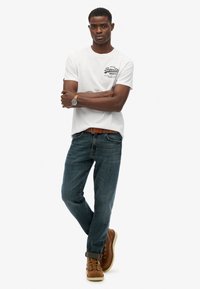 Camiseta blanca de manga corta con estampado gráfico negro, combinada con jeans de mezclilla azul oscuro y botas marrones con cordones y puños enrollados.