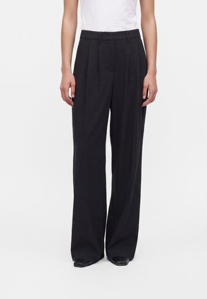 Persona che indossa pantaloni neri a vita alta, a gamba larga e con pieghe, abbinati a una camicia bianca infilata e scarpe nere, in piedi davanti a uno sfondo neutro.