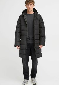 Melna pūkainā jaka ar kapuci, quilted dizains, garas piedurknes, priekšējais rāvējslēdzis un sānu kabatas, valkāta virs tumšas T-kreklā un melnām biksēm.