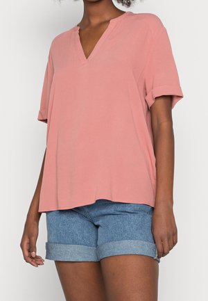 Rosa Kurzarm-Shirt mit V-Ausschnitt und aufgerollten Ärmeln, aus glattem Stoff gefertigt. Getragen mit Jeansshorts mit umgeschlagenem Saum.