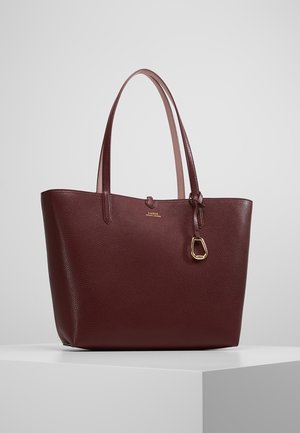 Ciemnobrązowa skórzana torba typu tote z podwójnymi paskami na ramię, złotym zawieszkiem z logo oraz subtelnym wytłoczonym napisem marki na przodzie.