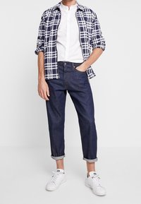 Niebiesko-biały koszula w kratę na białej koszuli z guzikami, w połączeniu z ciemnoniebieskimi dżinsami i białymi trampkami. Podwinięte mankiety spodni.