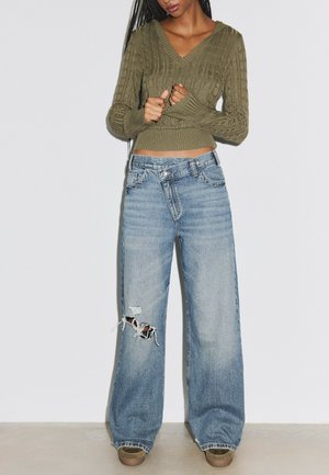 Personne portant un pull torsadé vert olive et un jean déchiré bleu clair ample avec des chaussures marron, debout contre un mur blanc.