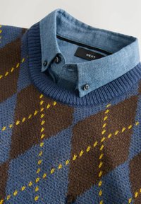 Der gestrickte Pullover hat ein braunes, blaues und gelbes Argyle-Muster, darunter ein Jeanshemd mit Kragen und gerippte blaue Bündchen.