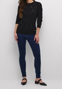 Schwarzer gestrickter Pullover mit Rautenmuster, rundem Ausschnitt und gerippten Säumen, kombiniert mit dunkelblauen Skinny-Jeans und schwarzen Blockabsatzschuhen.