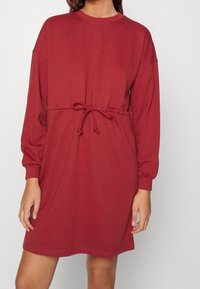 Robe rouge oversize en tissu doux, avec manches longues, encolure ronde et taille ajustable grâce à un cordon de serrage.