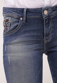 Jeans de mezclilla en un tono azul desvaído. Las características incluyen un bolsillo con cremallera en el frente, cierre de botón y un sutil desgaste en la tela.