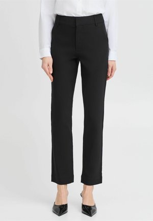 Femme portant un pantalon noir slim, une chemise blanche à manches longues et des escarpins noirs à bout pointu, debout devant un fond uni.