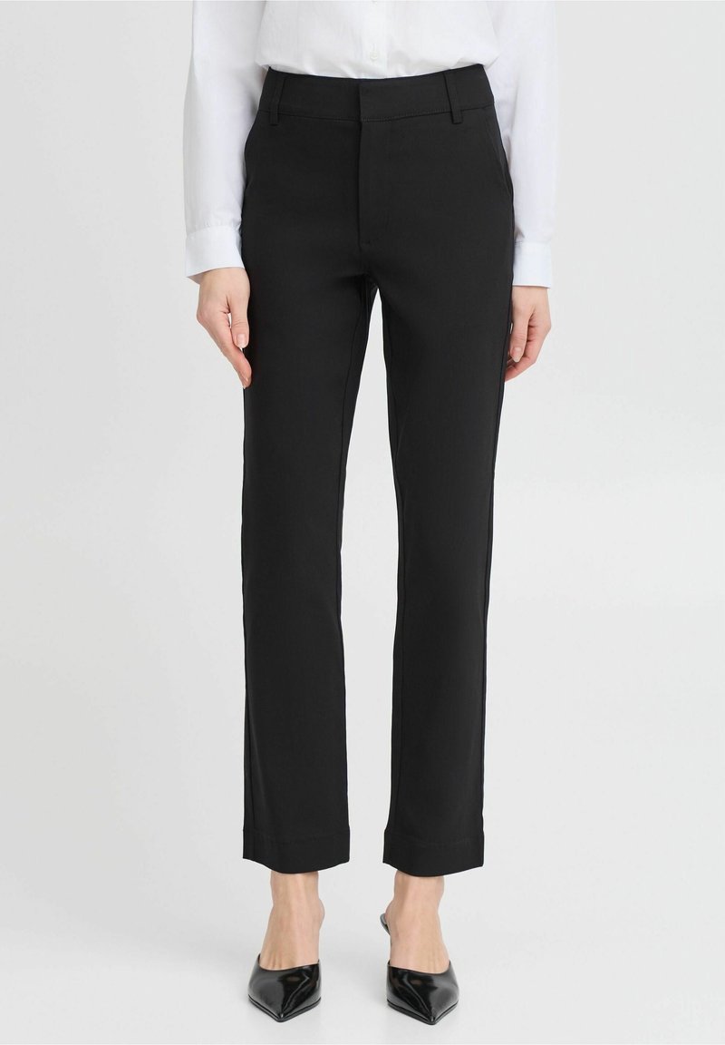 Femme portant un pantalon noir slim, une chemise blanche à manches longues et des escarpins noirs à bout pointu, debout devant un fond uni.