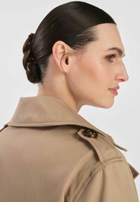 Beżowy trencz z wyraźnym kołnierzem, z detalem guzikowym na ramieniu. Gładka tekstura materiału. Włosy ułożone w schludny kok.