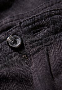 Donker stof met een visgraatpatroon, met een glanzende zwarte knop en een metalen rits die fijne stikdetails laat zien.