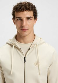 Beige dragkedjahoodie med svart dragkedja och snören. Har en sexkantig logotyp på bröstet och ett strukturerat tygmönster.