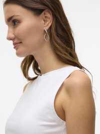 Jeune femme aux cheveux châtain clair, portant un haut blanc sans manches et des boucles d'oreilles argentées en boucle, tournée vers la gauche sur un fond uni.