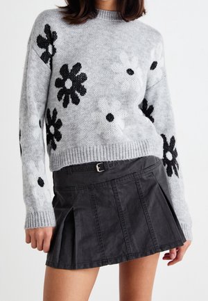 Femme portant un pull gris tricoté avec des motifs de fleurs noires et blanches et une mini-jupe plissée noire avec une ceinture.