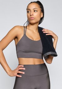 Sutiã de desporto cinza com tiras finas e design sem costuras, combinado com leggings de cintura alta a condizer. Uma toalha escura pendurada no ombro.