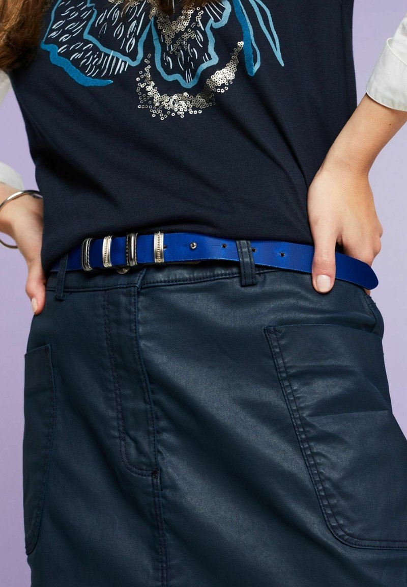 Personne portant un pantalon foncé avec de grandes poches, une ceinture bleue avec des anneaux argentés, et une chemise noire avec un motif floral bleu et argenté.