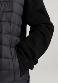 Gilet imbottito nero con maniche nere a coste testurizzate. Il gilet presenta pannelli orizzontali a cucitura stretta e una finitura liscia.